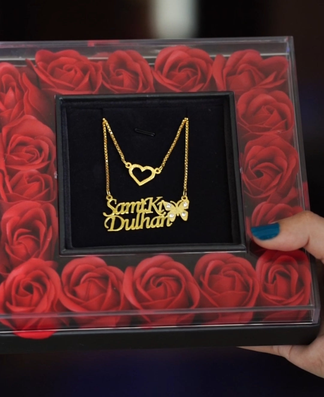 Gold necklace with 'Sam Ki Dulhan' on red roses in a box, held by a hand.