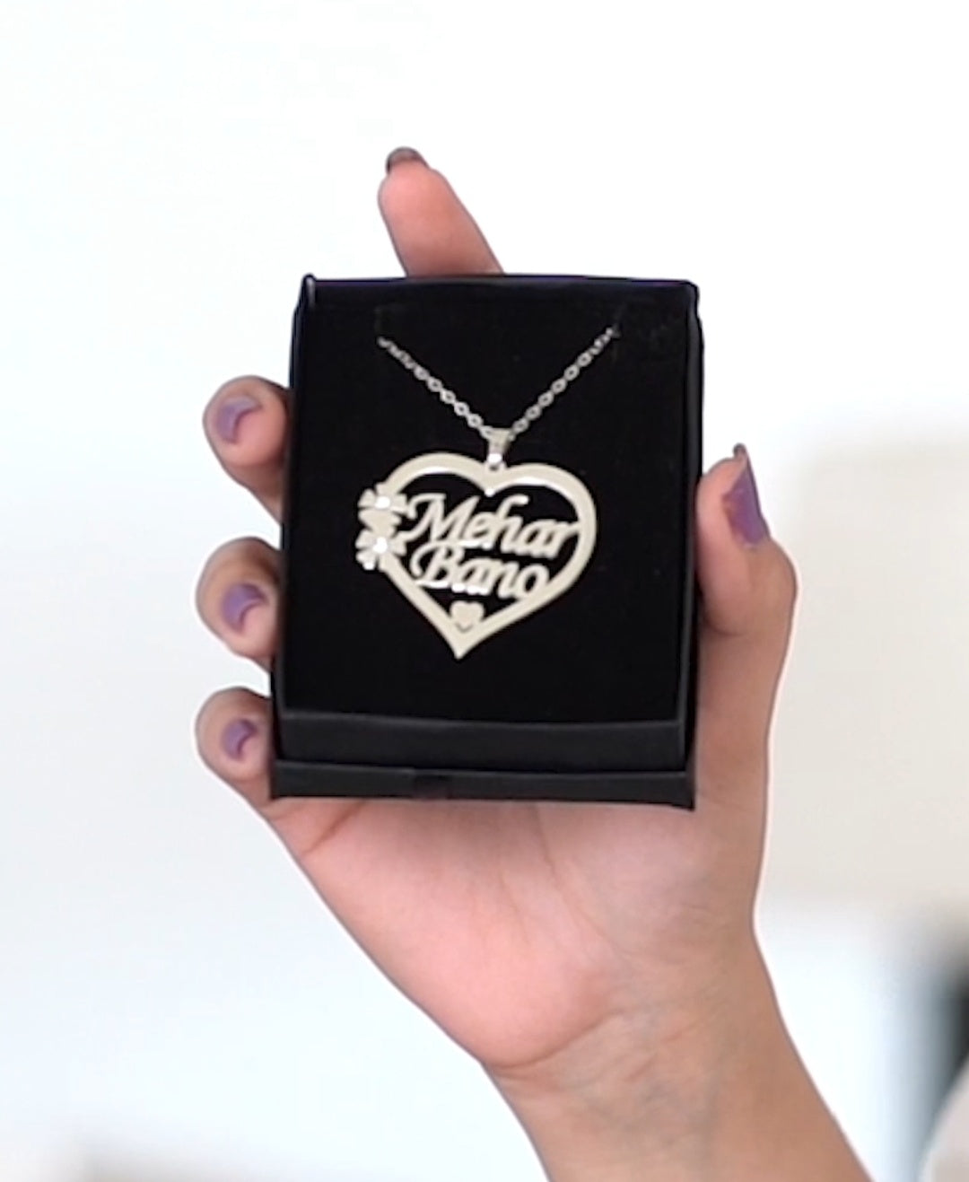 Heart-shaped necklace with 'Mehar Bano' text in a black box held by a hand.